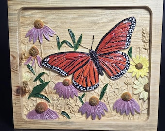 May include: A square wooden wall plaque featuring a relief carving of a large orange and black monarch butterfly surrounded by painted purple coneflowers and yellow daisies on a natural light wood grain background with green leaves.