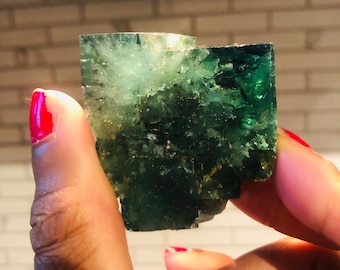 Könnte beinhalten: Ein grüner Fluorit-Kristall mit einer rauen, strukturierten Oberfläche. Der Kristall ist durchscheinend und hat einen leichten Schimmer.