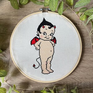 May include: A cross-stitch embroidery hoop with a white background and a red-winged, devil-themed cherub with a tail. The cherub has black hair and a light skin tone.