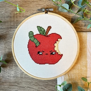 May include: A red apple with a bite taken out of it, a green worm inside, and a cute cartoon face. The apple is embroidered on a white canvas in a wooden embroidery hoop.