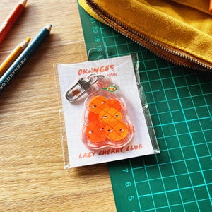 May include: A clear acrylic keychain with a design of orange cartoon oranges with sleepy eyes, on a lined paper card. The card reads "ORANGES" and "LAZY CHERRY CLUB". A silver-coloured clasp is attached.