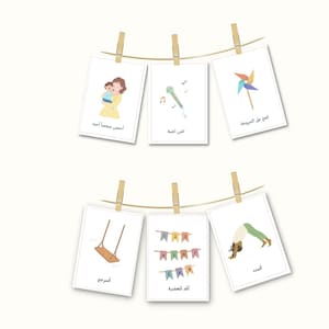 Könnte beinhalten: Sechs weiße Karten mit bunten Illustrationen und arabischem Text, die an einer Schnur mit Wäscheklammern hängen. Die Karten zeigen eine Mutter, die ein Kind hält, ein Mikrofon, ein Windrad, eine Schaukel, eine Girlande mit Flaggen und eine Person, die Yoga macht.