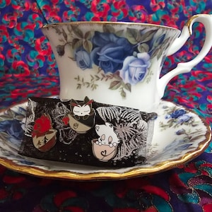 Puede incluir: Tres pines de esmalte con gatos de dibujos animados en tazas de té. Los pines están sobre un fondo de terciopelo negro y se muestran en una taza de té y un platillo blancos con un patrón floral.