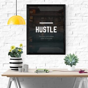 Puede incluir: Un póster motivacional enmarcado en negro con la palabra "Hustle" en texto blanco y la cita "The only controllable pillar of success" en una pared de ladrillo blanco con una lámpara amarilla colgando arriba y una planta en maceta sobre una mesa de madera.