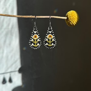 Peut inclure: Une paire de boucles d'oreilles en forme de goutte perlées. Les boucles d'oreilles présentent un motif noir, blanc, jaune et orange. Elles sont suspendues à des crochets argentés. Le fond est gris foncé avec une tige de fleur séchée.