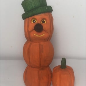 Puede incluir: Una figurita de calabaza de madera tallada con un sombrero verde y una cara sonriente. La calabaza está apilada sobre otras dos calabazas. Una calabaza más pequeña está a la derecha de la figurita.
