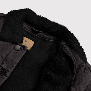 May include: Close-up of a dark gray denim jacket with a black, fluffy shearling collar and lining. The jacket has a button-down front and a small tag that reads "M". The jacket is a medium size.