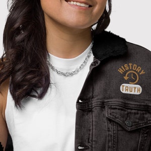 May include: A black denim jacket with a black faux fur collar. The jacket features a patch with the words "HISTORY" and "TRUTH" surrounding a clock and arrow design. A silver chain necklace is also visible.