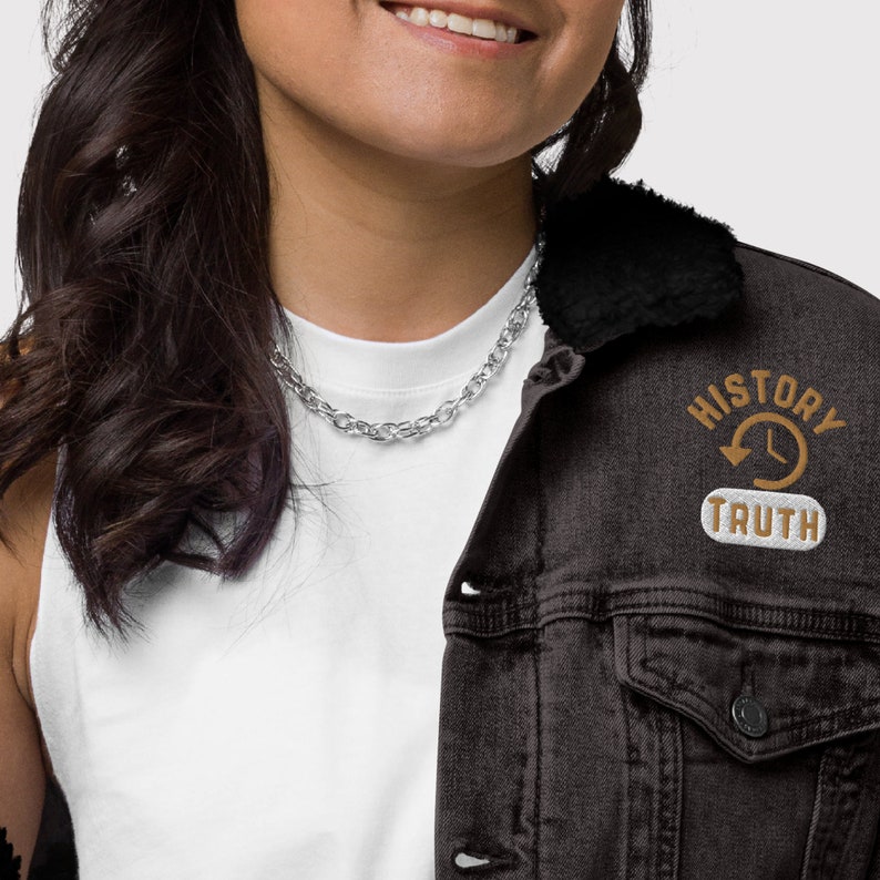 May include: A black denim jacket with a black faux fur collar. The jacket features a patch with the words "HISTORY" and "TRUTH" surrounding a clock and arrow design. A silver chain necklace is also visible.