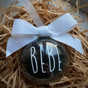 May include: A black and silver glitter Christmas ornament with the word "BEBE" in white lettering. The ornament is topped with a white ribbon bow and is nestled in brown paper filler.