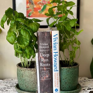 May include: Two green ceramic pots containing plants, one with basil and the other with mint, are used as bookends for three books. The books are "This Will Make It Taste Good" and "Deep Run Roots" by Vivian Howard.