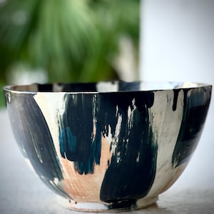 May include: A ceramic bowl with a hand-painted design. The bowl features a white base with bold brushstrokes of black, teal, and peach. The interior is white, and the rim has a dark, dripped paint effect. This decorative bowl is suitable for serving.