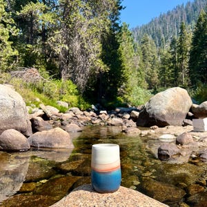 May include: A ceramic mug with a white, orange, and blue glaze sits on a rock by a stream in a forest setting.