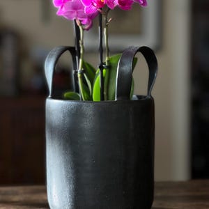 May include: A black, cylindrical planter with two arched handles, holding a vibrant pink orchid. The orchid has multiple blooms and green leaves. The planter has a textured surface and a rounded base.