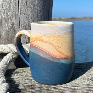 Può includere: Una tazza in ceramica con smalto multicolore che presenta una transizione graduale da una base blu navy profondo a una parte superiore color rosa tenue e beige sabbia. La tazza è dotata di un manico curvo e un corpo cilindrico con bordo.