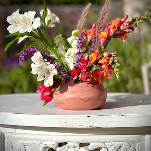 May include: A bouquet of white, pink, and orange flowers in a terracotta-colored ceramic vase. The vase is sitting on a white table with intricate carvings.