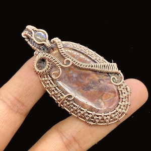 May include: A handcrafted pendant featuring a polished, oval-shaped stone with a mix of brown, red, and cream colors. The stone is intricately wrapped in copper wire, creating a decorative frame. A small, iridescent stone is set at the top.