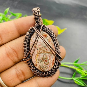 May include: A copper wire wrapped pendant featuring a brown, white, and pink stone with a swirling pattern. The pendant is oval shaped and has a woven wire design.