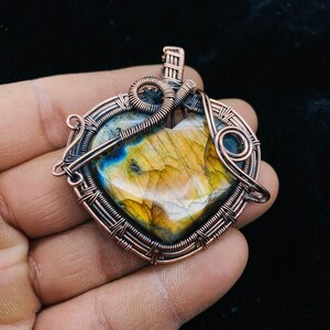 May include: A copper wire wrapped heart-shaped pendant featuring a large iridescent labradorite gemstone in the centre. The pendant has a copper bail for attaching to a chain.