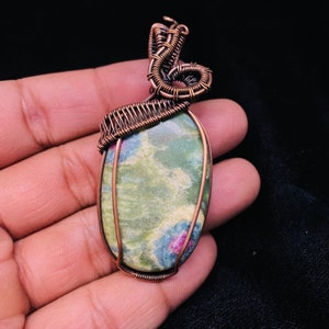 Puede incluir: Un colgante de piedra preciosa verde y rosa envuelto en alambre de cobre. El colgante tiene forma ovalada y presenta un patrón único de remolino.