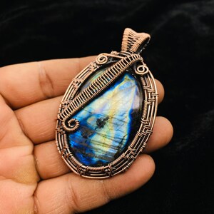 May include: A copper wire wrapped pendant featuring a blue and green labradorite gemstone. The pendant has a woven wire design and a loop at the top for attaching a chain.