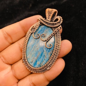 Könnte beinhalten: Handgefertigter Anhänger aus Kupferdraht mit einem blauen Edelstein. Der oval geformte Stein hat eine hellblaue Farbe mit dunkleren blauen Streifen. Der Kupferdraht ist kunstvoll um den Stein gewebt.