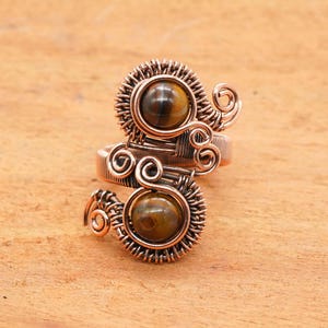 May include: Copper wire-wrapped ring featuring two round, polished tiger's eye stones. The ring has an open design with intricate wirework around the stones and on the band. The ring is set against a light brown wooden surface.