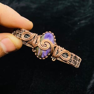 Pode incluir: Uma pulseira artesanal de fio de cobre com uma pedra central oval roxa e verde. O fio é intrincadamente tecido ao redor da pedra e ao longo da pulseira, criando um design artístico único. A pulseira é segurada por uma mão contra um fundo escuro.