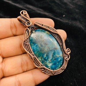 Puede incluir: Un colgante envuelto en alambre de cobre con una piedra preciosa ovalada azul y verde. El colgante tiene un diseño de alambre detallado con un remolino en la parte superior.
