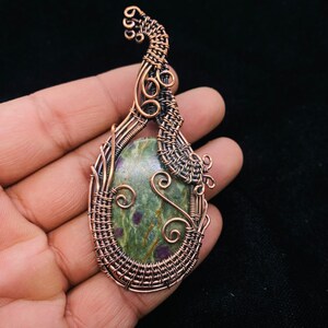 May include: A copper wire wrapped pendant featuring a green and pink gemstone. The wire is woven into a delicate, intricate design.
