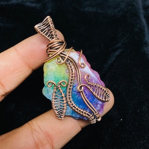 Op de afbeelding: Een regenboog-agaten hanger gewikkeld in koperdraad. De draad is gedraaid en gevormd tot een delicaat ontwerp. De agaat is een natuursteen met een verscheidenheid aan kleuren, waaronder roze, paars, groen en geel.
