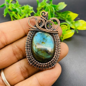 May include: A copper wire wrapped pendant featuring a green and blue labradorite gemstone. The pendant has a unique design with a woven wire frame and a decorative swirl at the top.