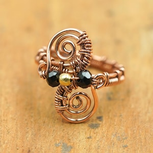 May include: A handcrafted copper wire ring featuring a unique design with spiral accents. The ring is adorned with black and gold beads, adding contrast to the copper color. This piece of jewelry is a statement accessory.