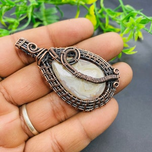 May include: A white agate pendant with a copper wire wrap. The wire wrap is intricate and features a woven design. The pendant is a teardrop shape and is perfect for adding a touch of natural beauty to any outfit.