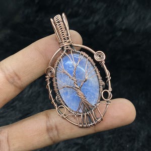 Op de afbeelding: Een hanger van koperdraad met een blauwe edelsteen met een boomontwerp. De hanger is ovaalvormig en heeft een koperen lus om op te hangen.