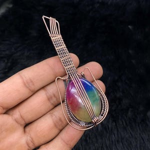 Op de afbeelding: Een gitaar hanger gemaakt van koperdraad met een regenboog agaat steen. De hanger is een uniek en kleurrijk sieraad.