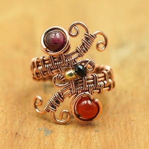 May include: A copper wire ring with an artistic design. The ring features a deep red gemstone, a black gemstone, and a golden bead. The intricate wirework and gemstone accents create a distinctive piece of jewellery. The ring is a unique, eye-catching piece.