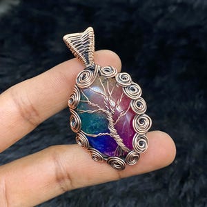 Op de afbeelding: Een hanger van koperdraad gewikkeld rond een veelkleurige agaat-steen met een levensboom-ontwerp. De steen is een mix van roze, groen, blauw en paars.