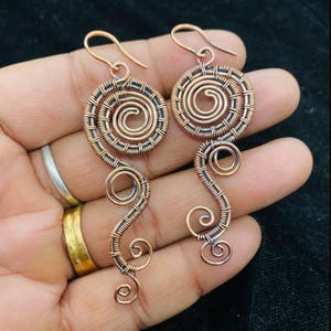 May include: Copper wire earrings with a spiral design. The earrings feature a circular top with a central spiral and a flowing, curved bottom section. The copper color is visible throughout the design, with a hook for the ear.