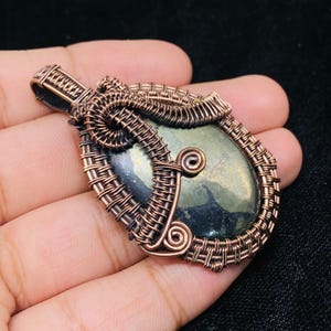 May include: A handcrafted pendant featuring a polished, oval-shaped stone in shades of green and gray, intricately wrapped with copper wire. The wirework includes woven patterns and decorative spirals, creating a unique, artistic design.