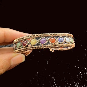 May include: A copper-colored wire-wrapped bracelet featuring a row of colorful gemstones. The bracelet has a black band in the center and is held by a hand against a dark background. The gemstones include green, red, yellow, purple, orange, blue, and white.