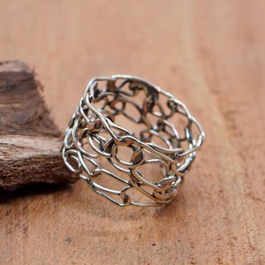 May include: A silver ring with an open, woven design. The ring's intricate pattern features interconnected loops and lines, creating a textured appearance. The ring is displayed on a wooden surface, showcasing its metallic sheen.