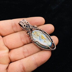 Könnte beinhalten: Ein mit Kupferdraht umwickelter Anhänger mit einem blauen und goldenen Labradorit-Edelstein. Der Anhänger ist oval geformt und hat ein detailliertes Drahtdesign.