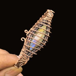 Puede incluir: Una pulsera artesanal de alambre de cobre con una piedra central de labradorita iridiscente. La piedra está encerrada en un diseño en forma de jaula, con un intrincado enrollado de alambre. La pulsera tiene un tono cálido y terroso.