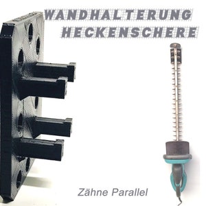 Puede incluir: Un soporte de pared de plástico negro con tres ranuras para sujetar cortasetos. El texto "WANDHALTERUNG HECKENSCHEERE" está impreso en blanco en el soporte de pared. El texto "Zähne Parallel" está impreso en negro debajo del soporte de pared. Un cortasetos se muestra colgado en el soporte de pared.
