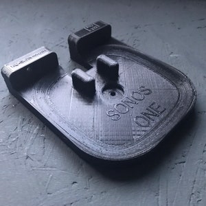 Peut inclure: Un support mural imprimé en 3D noir pour une enceinte Sonos One. Le support comporte deux languettes à l'arrière pour le montage mural et le texte "SONOS ONE" est imprimé à l'avant.