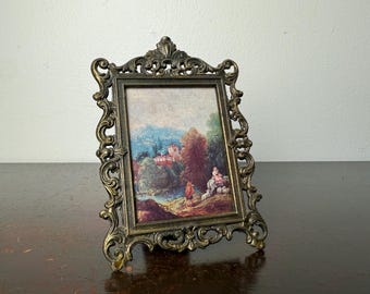Puede incluir: Marco de fotos antiguo de latón ornamentado con una pintura de paisaje. La pintura representa una escena con un río, árboles y un edificio. El marco tiene un diseño detallado y en espiral, sobre una superficie de madera.