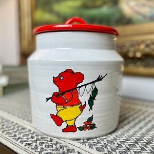 Puede incluir: Tarro de galletas de cerámica blanca con tapa roja. El tarro presenta una ilustración de dibujos animados de un oso rojo con pantalones amarillos y sosteniendo una caña de pescar.