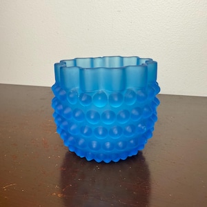 May include: A blue hobnail glass votive candle holder. The round, frosted glass container has a ruffled top edge and a textured surface of small, raised bumps. The color is a bright, vibrant blue.