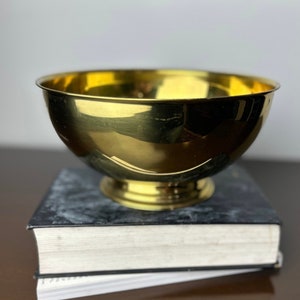 May include: A shiny gold metal bowl with a rounded bottom and a wide rim. The bowl is sitting on top of a black book.